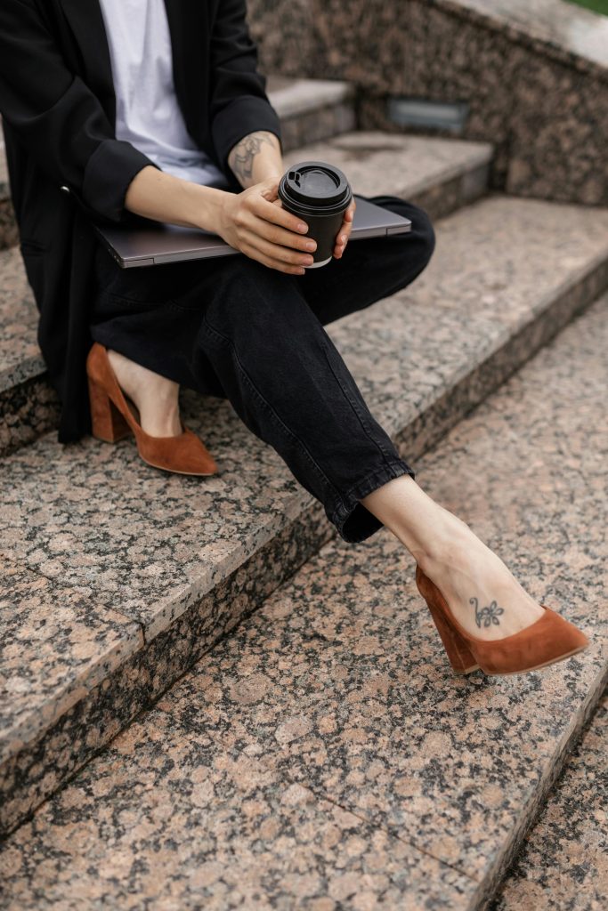 Stylish woman in heels enjoying coffee on stone steps, holding laptop.