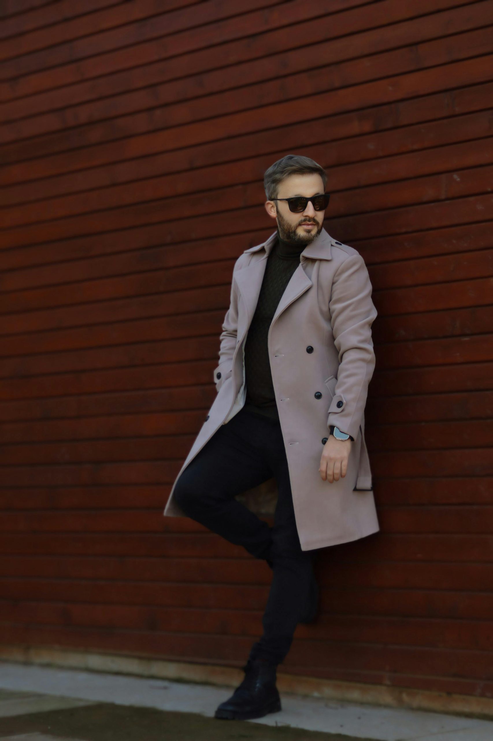 Stylish man wearing a trench coat and sunglasses against a wooden wall.